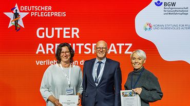 Jörg Schudmann, Hauptgeschäftsführer der BGW, zusammen mit Marita Frerichs und Stefanie Freimuth-Hunfeld, die die Urkunde hält, vom St. Nikolausstift Caritas Pflege aus Papenburg auf der Bühne. .