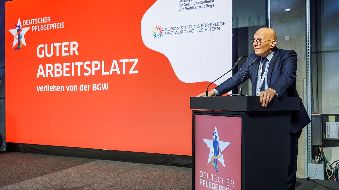 Person mit Brille, die ihre Hände auf ein Rednerpult stützt, links von der Person ein Leuchtdisplay mit Aufschrift.