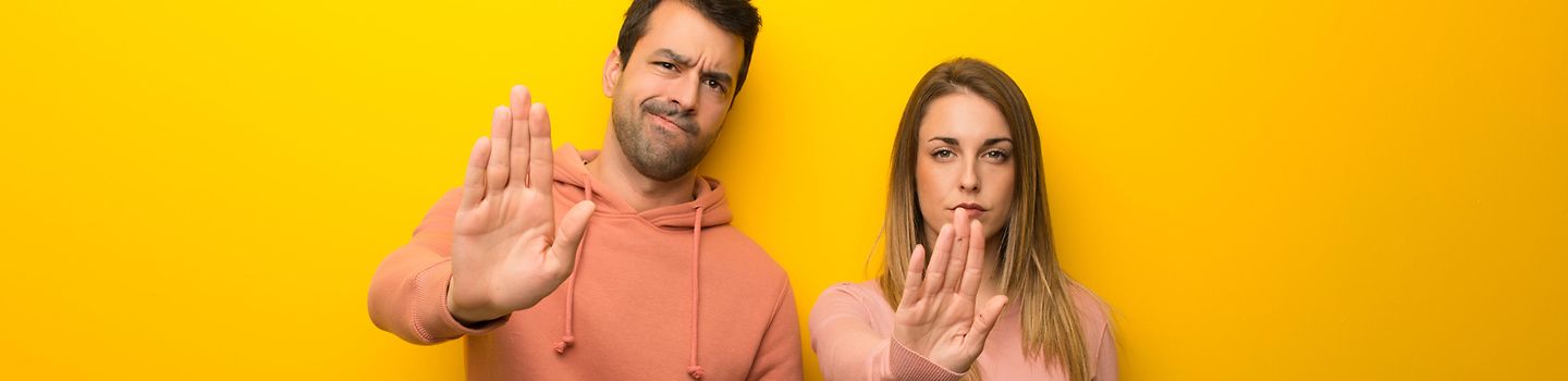 Zwei Personen vor gelbem Hintergrund. Beide zeigen eine Stop-Geste mit der rechten Hand. Ihr Gesichtsausdruck ist abwehrend.