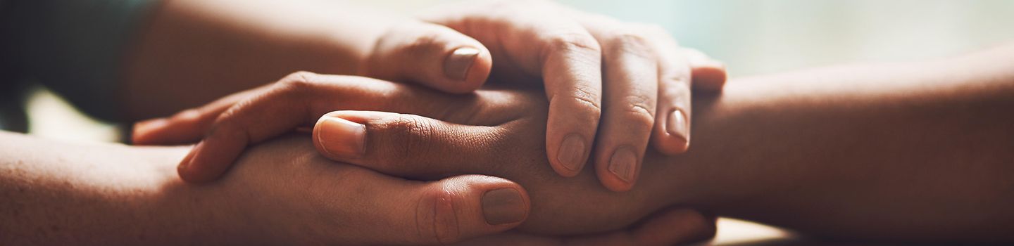 Love, empathy and support with people holding hands in comfort, care or understanding on a wooden table of a home. Prayer, faith or depression with friends closeup for hep, hope or peace for healing Eine Person hält die Hand einer anderen zur Unterstützung. Die Hände liegen auf einem Tisch ab.