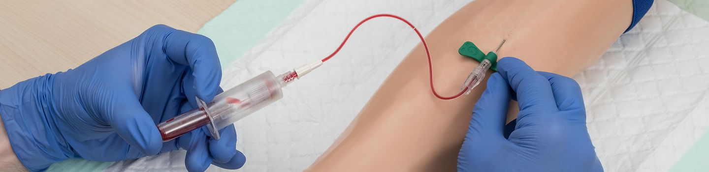Zwei Hände in blauen Handschuhen ziehen Blut aus einem künstlichen Übungsarm durch eine Kanüle in ein Blutentnahmesystem auf.