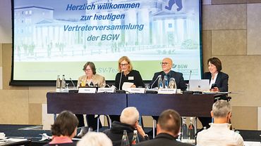 Vier Personen sitzen in einer Reihe nebeneinander auf einer Bühne, vor ihnen Publikum