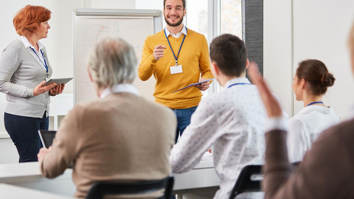 Ein lächelnder Mann steht vor einer Gruppe von Menschen in einem Seminar neben einem Whiteboard. Ein lächelnder Mann steht vor einer Gruppe von Menschen in einem Seminar neben einem Whiteboard.
