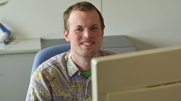 Yannick Ludewig BGW-Mitarbeiter Yannick Ludewig sitzt am Schreibtisch vor einem Monitor und lächelt in die Kamera.