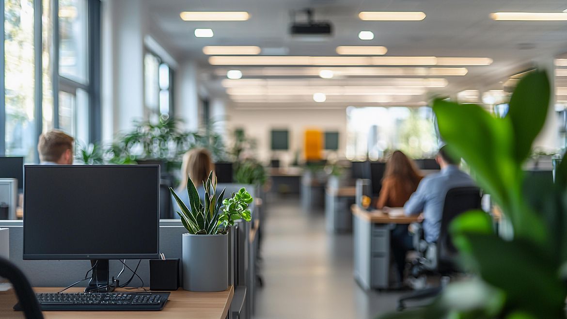 Modern eingerichtetes Großraumbüro mit mehrern Reihen mit Schreibtischen und Bildschirmarbeitzplätzen. Im Vordergrund eine Grünpflanze, der vorderste Arbeitsplatz ist leer, dahinter vier Männer und Frauen am Schreibtisch