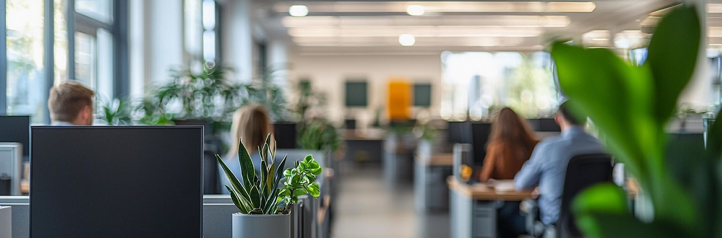 Großraumbüro Modern eingerichtetes Großraumbüro mit mehrern Reihen mit Schreibtischen und Bildschirmarbeitzplätzen. Im Vordergrund eine Grünpflanze, der vorderste Arbeitsplatz ist leer, dahinter vier Männer und Frauen am Schreibtisch