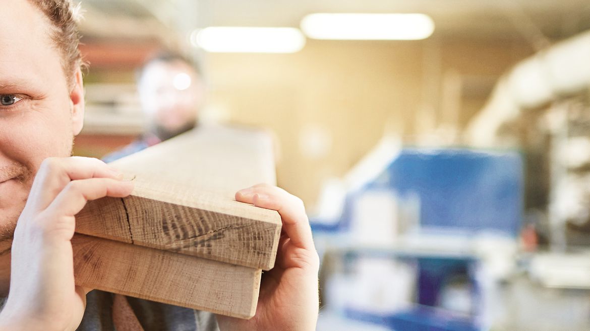 Zwei Männer in einer Holzwerkstatt tragen zwei lange, dicke und schwere Bretter auf ihren Schultern