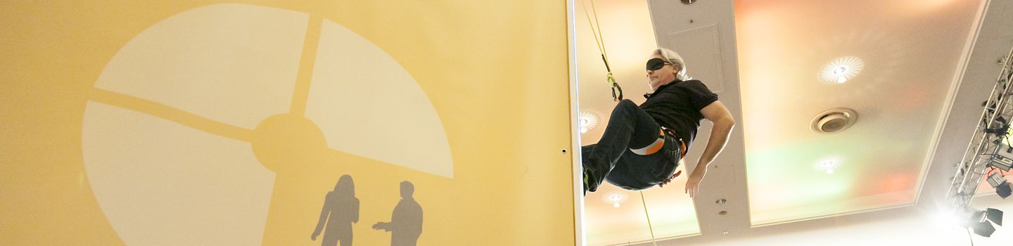 Mann in Kletterausrüstung und mit einer Augenbinde klettert seitlich an einer Wand, die mit einem Logo bedruckt ist, das drei Kreissegmente und zwei Figuren zeigt.