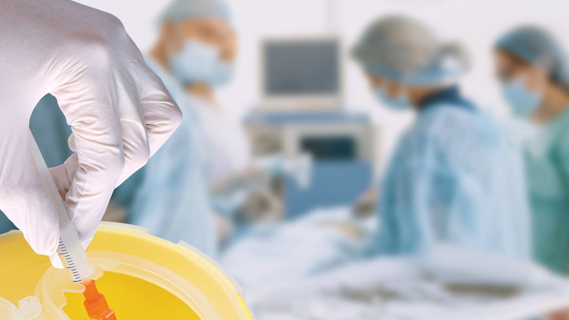 Eine Hand in medizinischem Handschuh hält eine Spritze. Die von einer Schutzkappe verdeckte Kanüle wird in einen Behälter abgestreift. Im Hintergrund, unscharf, ein OP-Team beim Operieren.