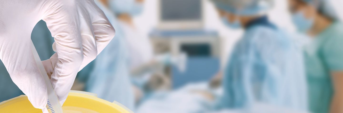 Eine Hand in medizinischem Handschuh hält eine Spritze. Die von einer Schutzkappe verdeckte Kanüle wird in einen Behälter abgestreift. Im Hintergrund, unscharf, ein OP-Team beim Operieren.