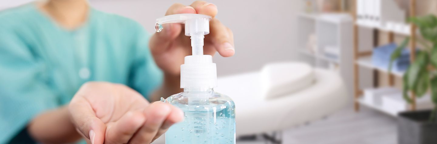Nahaufnahme eines Gelspenders in einer Massagepraxis. Eine Frau in mintgrünem Kittel betätigt den Spender mit einer Hand, die andere Handfläche geöffntet darunter. Im Hintergrund eine weiße Behandlungsliege im Praxisraum.
