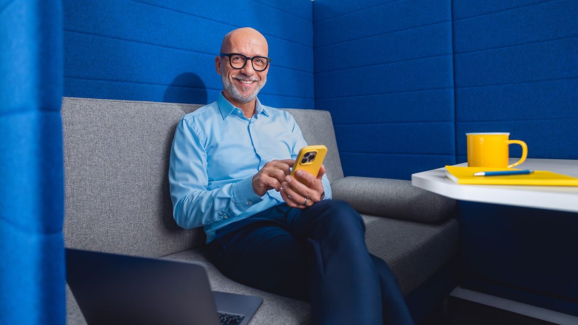 1000UNDEIN GRUND Ein Mann sitzt auf einem Sofa vor einer blauen Wand und hält ein Smartphone in der Hand.