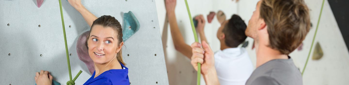 Frau klettert eine Kletterwand hoch und wird von einem Mann gesichert.