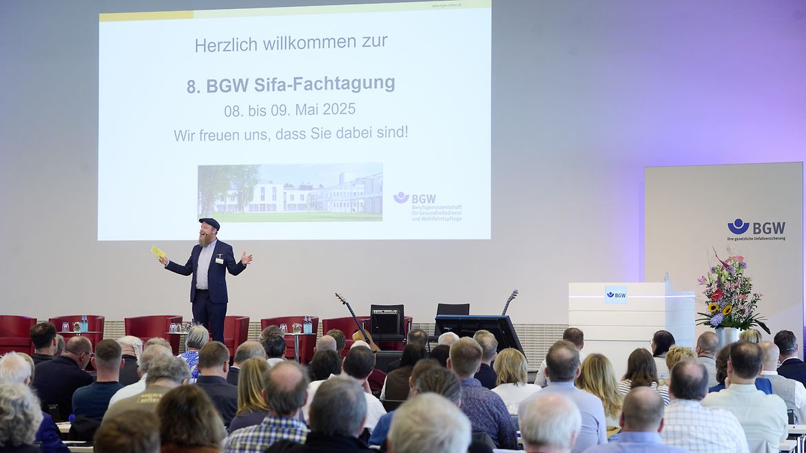 Ein Mann mit Bart und Mütze steht vor Publikum auf einer Bühne, auf der Bühne hinter ihm steht per Beamer "Herzlich willkommen zur 8. BGW Sifa-Fachtagung"