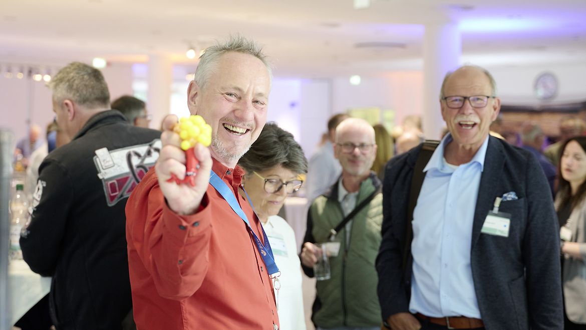 Zwei Männer und eine Frau stehen gemeinsam im Foyer und lachen, ein Mann hält einen bunten Gegenstand in die Kamera.