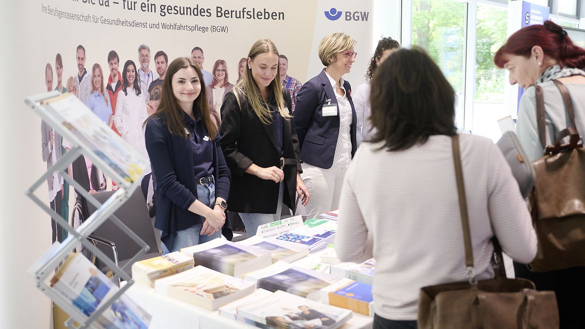 Vier Frauen stehen hinter einem Info-Stand mit Broschüren, davor sehen sich zwei Frauen die Materialien an.