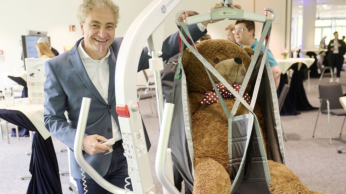 Ein Mann bedient einen Patientenlifter, in dem ein sehr großer Teddy-Bär sitzt