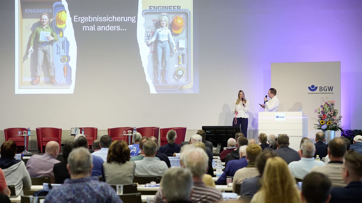 Ein Mann und eine Frau stehen auf der Bühne der Sifa-Fachtagung, an die Wand projeziert steht "Ergebnissicherung mal anders" und es sind "Actionfiguren" von einer Ingenieurin und einem Ingenieur zu sehen.