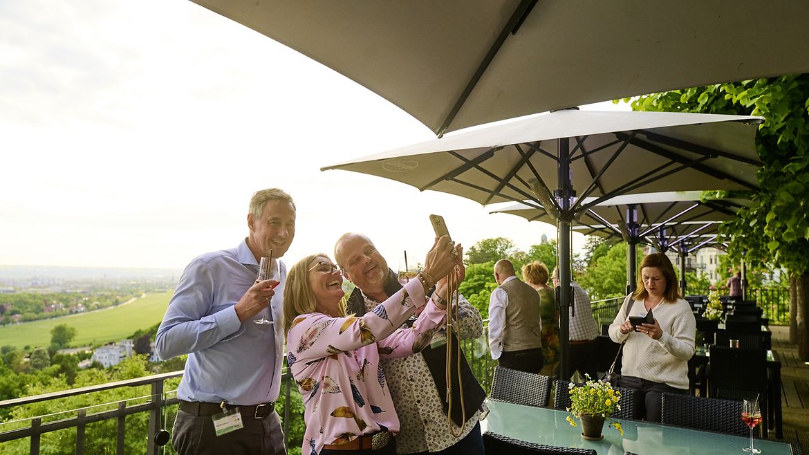 Zwei Männer und eine Frau stehen auf einer Terasse mit Getränken in der Hand, die Frau macht ein Selfie der Gruppe. Im Hintergrund ist weite, grüne Landschaft zu sehen.