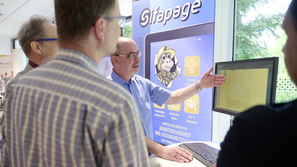 Ein Mann zeigt drei anderen Männern etwas auf einem Computermonitor, dahinter steht ein Aufsteller mit dem Wort "Sifapage".