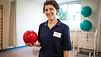 Eine Physiotherapeutin steht in einem Reha-Raum und hält einen kleinen Medizinball in der Hand