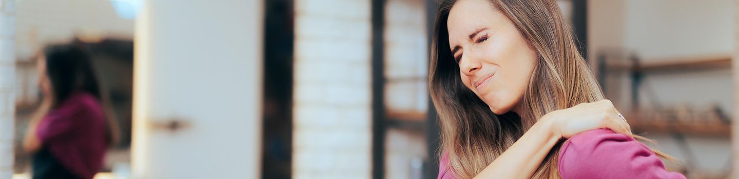 Friseurhandwerk Junge Friseurin hält sich mit der rechten Hand die linke Schulter und kneift das Gesicht zusammen