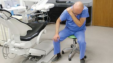 Schulterschmerz in der Zahnarztpraxis Mann, der auf einem Drehhocker sitzt, sich vornüber beugt und die Schulter hält. Hinter ihm medizinische Geräte.