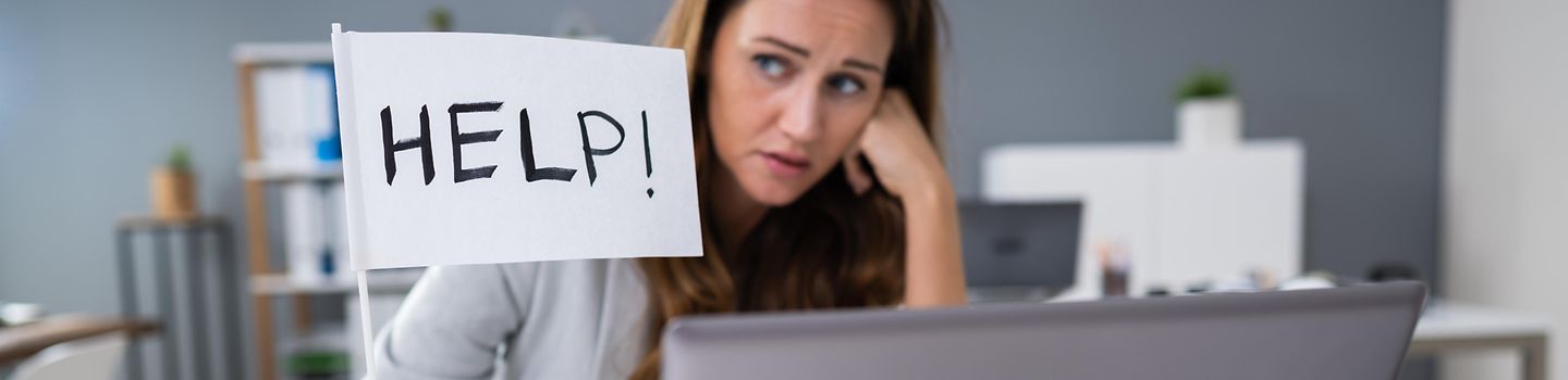 Unglücklich aussehende Frau sitzt im Büro am Schreibisch vor einem aufgeklappten Laptop mit einem Fähnchen in der Hand, auf dem Help! steht