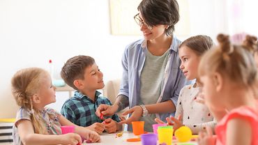 Erzieherin mit vier Kindern an einem Tisch mit bunten Bechern, Deckeln und Knetmasse
