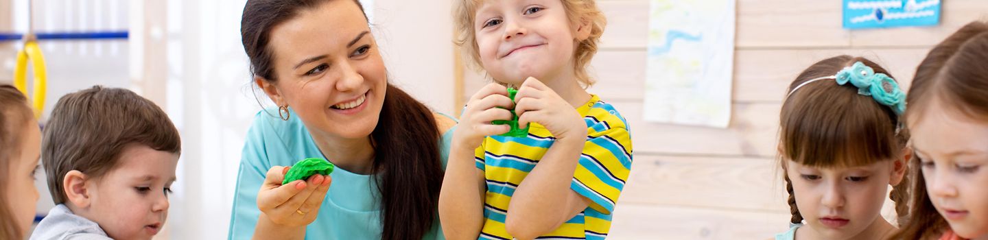 Fünf Kleinkinder sitzen zusammen mit einer Betreuuerin an einem Tisch und spielen mit Knetgummi.