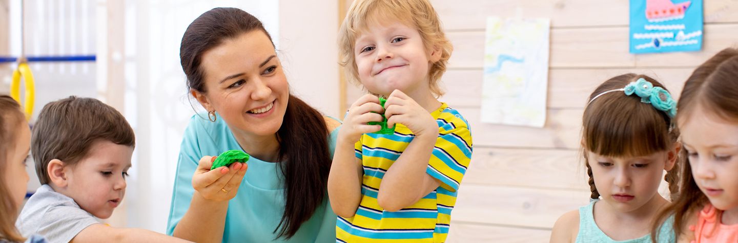 Fünf Kleinkinder sitzen zusammen mit einer Betreuuerin an einem Tisch und spielen mit Knetgummi.