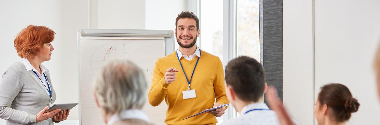 Ein lächelnder Mann steht vor einer Gruppe von Menschen in einem Seminar neben einem Whiteboard. Ein lächelnder Mann steht vor einer Gruppe von Menschen in einem Seminar neben einem Whiteboard.