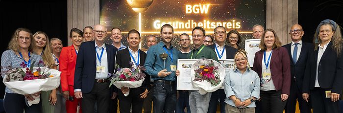 Gruppenbild der Finalteilnehmenden mit Jury-Mitgliedern und dem Hauptgeschäftsführer der BGW bei der Verleihung des BGW-Gesundheitspreises auf der Bühne.