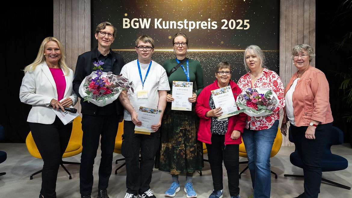 Die Preisträgerinnen und Preisträger Matthias Bostelmann (3.v.l.), Pia Grafunder (4.v.l.), Andrea Breitlow (5.v.l.) zusammen mit Betreuenden, Laudatorin Silvia Thimm (r.) aus der Selbstverwaltung der BGW und Moderatorin Katrin Degenhardt (l.) .