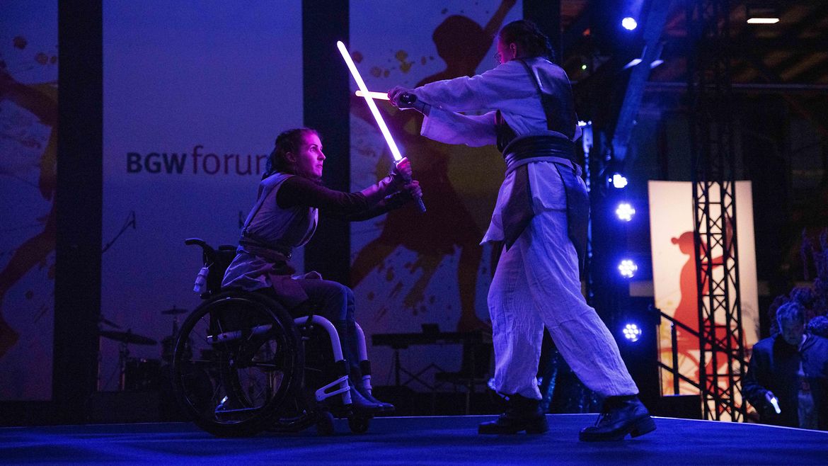 Zwei Personen, eine davon benutzt einen Rollstuhl, fechten mit Lichtschwertern