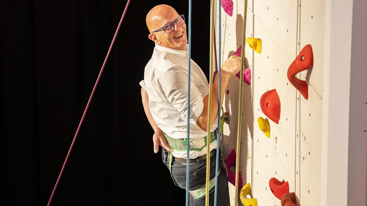 Eine Person am Kletterturm. Sie lächelt, eine Hand am Haltegriff, eine andere in der hinteren Hosentasche.