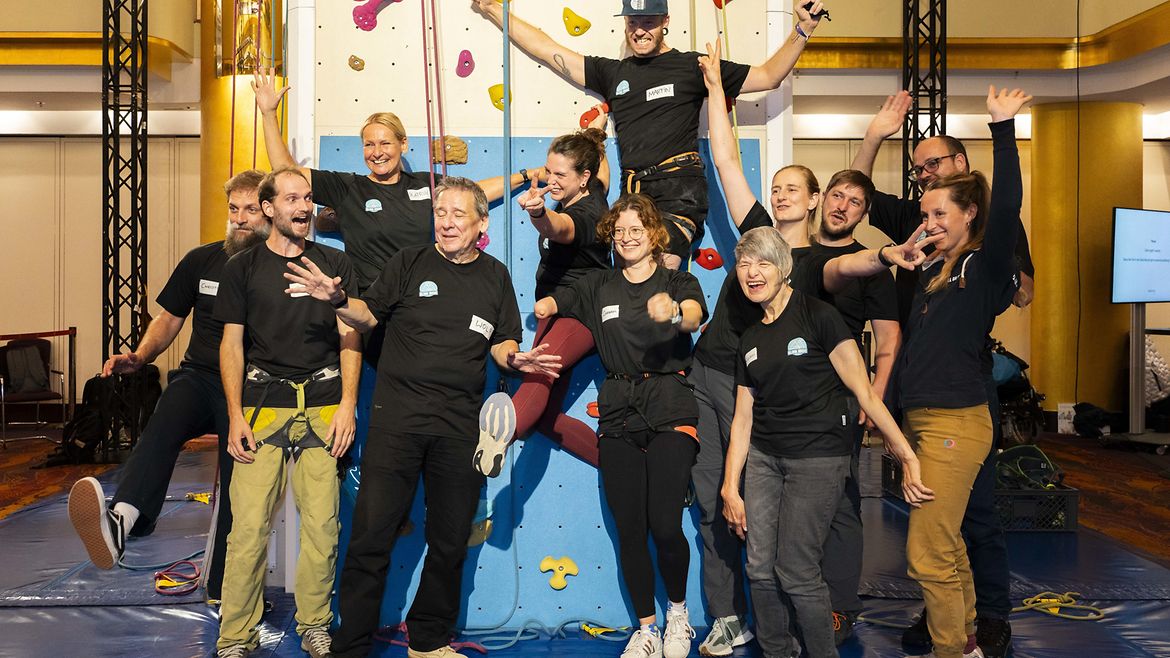 Zwölf Personen, platziert vor einem Kletterturm. Einige Personen hängen am Turm, einige stehen davor, einige haben die Hände zum Jubel erhoben, alle lächeln