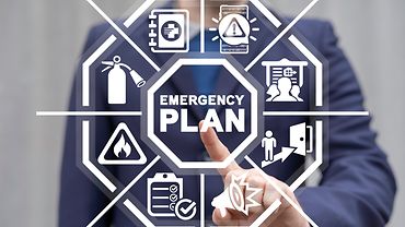Im Vordergrund eine weiße, sechseckige Grafik mit den Worten "Emergency Plan" in der Mitte und Notfallsymbolen wie Feuerlöscher drumherum. Im Hintergrund die Silhouette eines Mannes, dess Finger auf die Grafik zeigt.