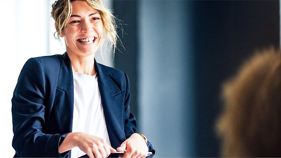 Eine junge Frau steht lächelt mit einem Tablet in der Hand, im Vordergrund sind Hinterköpfe von Personen zu erkennen, die sie anschauen.
