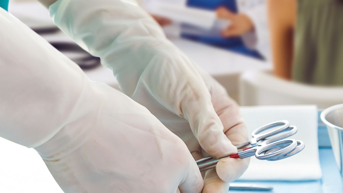 Im Hintergrund eine Ärztin im Gespräch mit Patientin. Im Vordergrund legt eine Person medizinische Instrumente für einen kleinen operativen Eingriff bereit.