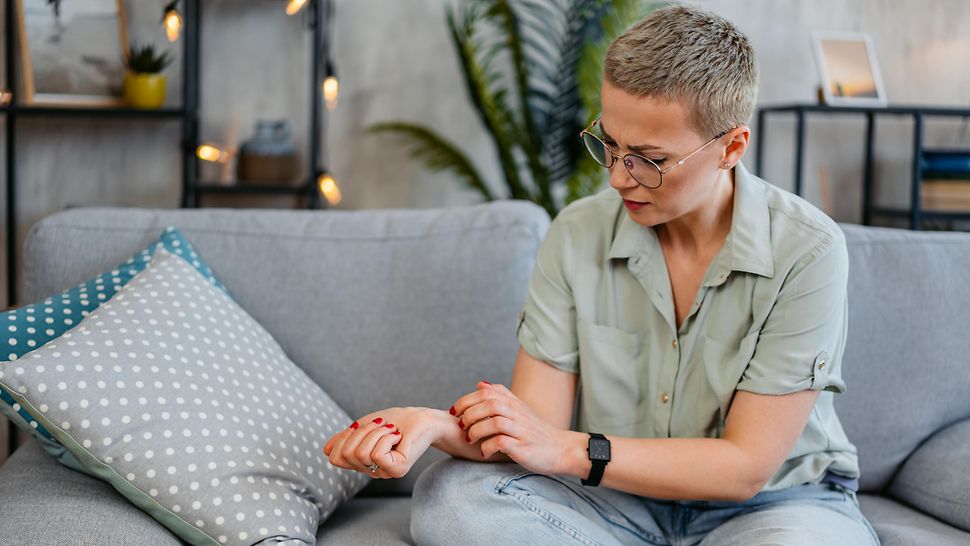 Eine Frau mittleren Alters in lockerer Pose auf einem Sofa. Sie schaut auf ihren rechten Arm, die Hand zur Faust geballt. Mit der anderen Hand kratzt sie eine Stelle unterhalb des Handgelenks