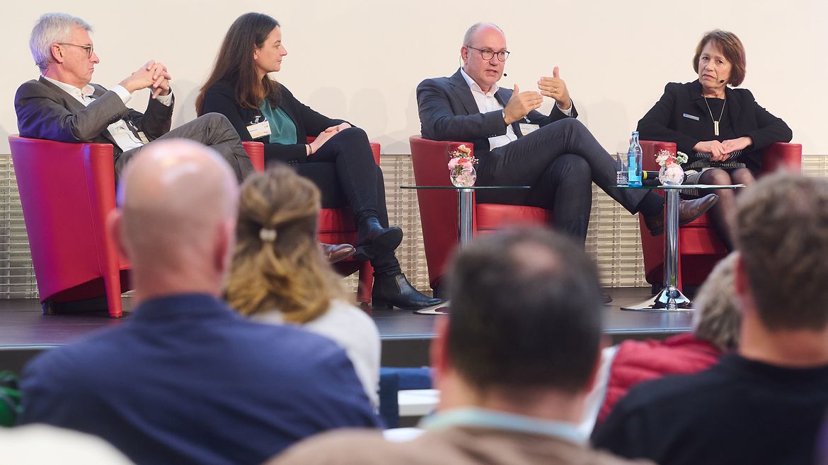 Blick aus dem Publikum auf eine Bühne, auf der zwei Mäönner und zwei Frauen in Sesseln sitzen und diskutieren.