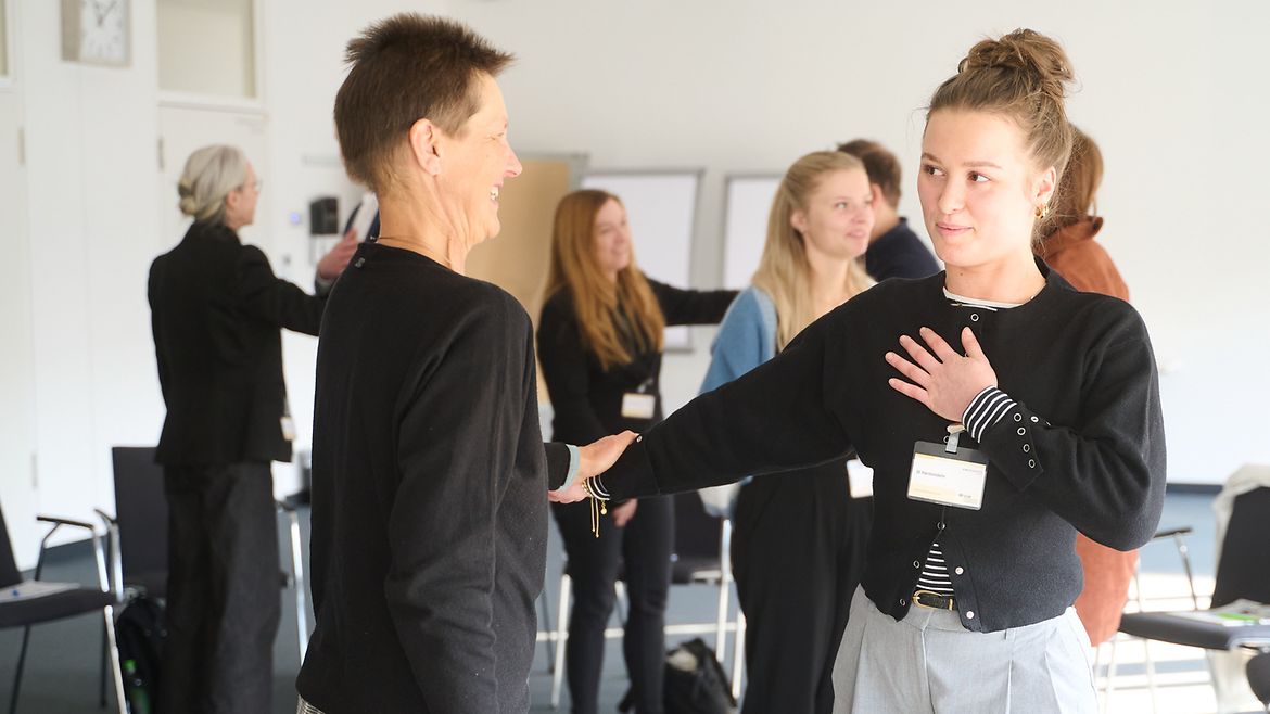 Zwei Frauen stehen sich bei einem Workshop gegenüber und halten sich am Arm fest, eine Frau hält die Hand auf ihre Brust.