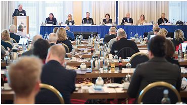 Ein Saal, an der Stirnseite ein Pult und ein langer Tisch an denen Personen sitzen, vor ihnen ein Publikum