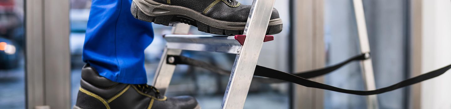 Person in Arbeitskleidung mit Sicherheitsschuhen. Die Person steigt eine Leiter hinauf.