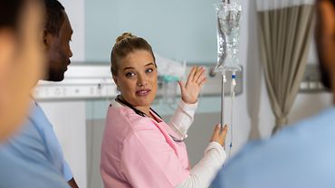 Weibliche Person weist mit den Händen in Richtung eines Infusionsständers. Sie blickt über ihre Schulter zurück auf eine weitere Person.