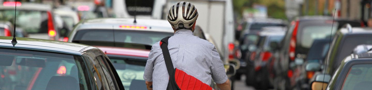 Rückansicht eines Fahrradfahrers mit Tasche und Helm zwischen vielen Autos, die in der Stadt vor einer grünen Ampel im Hintergrund stehen bzw. fahren