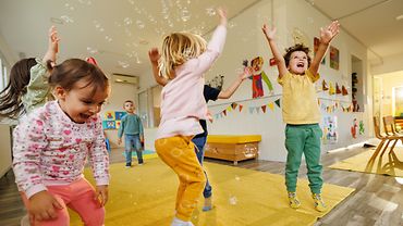 Jungen und Mädchen im Alter von etwa drei bis vier Jahren im Spielzimmer einer Kita. Sie springen hoch und lachen, über ihnen schweben Seifenblasen, die sie sie fangen