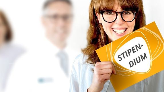 Junge Frau mit Brille hält ein Schild mit dem Wort "Stipendium" in die Kamera. Im Hintergrund drei Ärztinnen in weißen Mänteln.