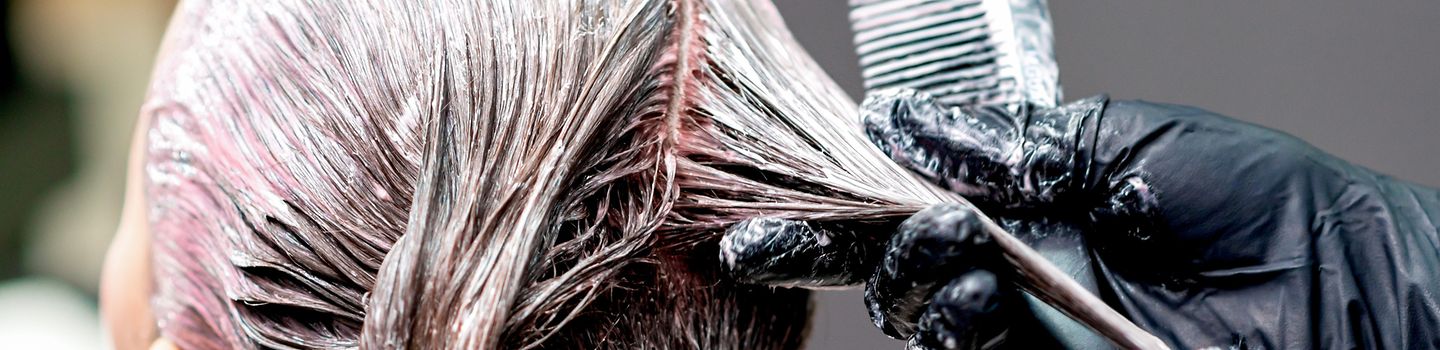 Friseurin oder Friseur beim Auftragen von Farbe auf den Kopf einer Kundin. Die Friseurin oder der Friseur trägt schwarze Handschuhe mit Farbspuren und hält einen Kamm in der Hand